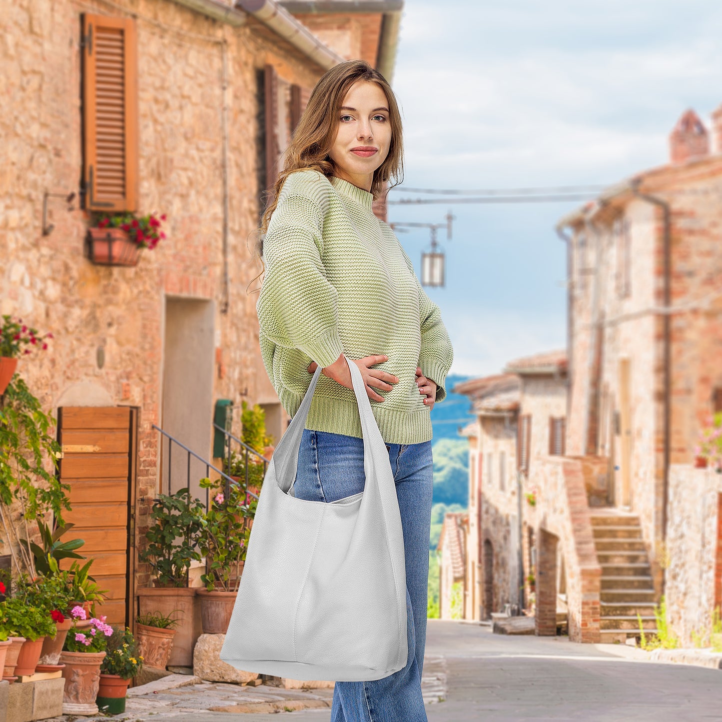 Woman in a light green sweater holds a white bag on a cobblestone street surrounded by buildings.
