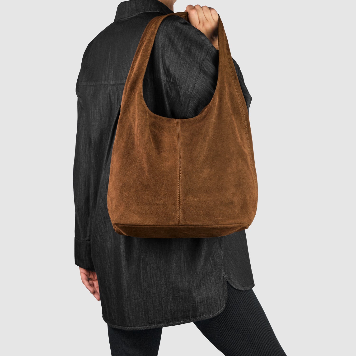Person wearing a black jacket and leggings, holding a large brown suede shoulder bag with a wide strap against a plain light gray background.