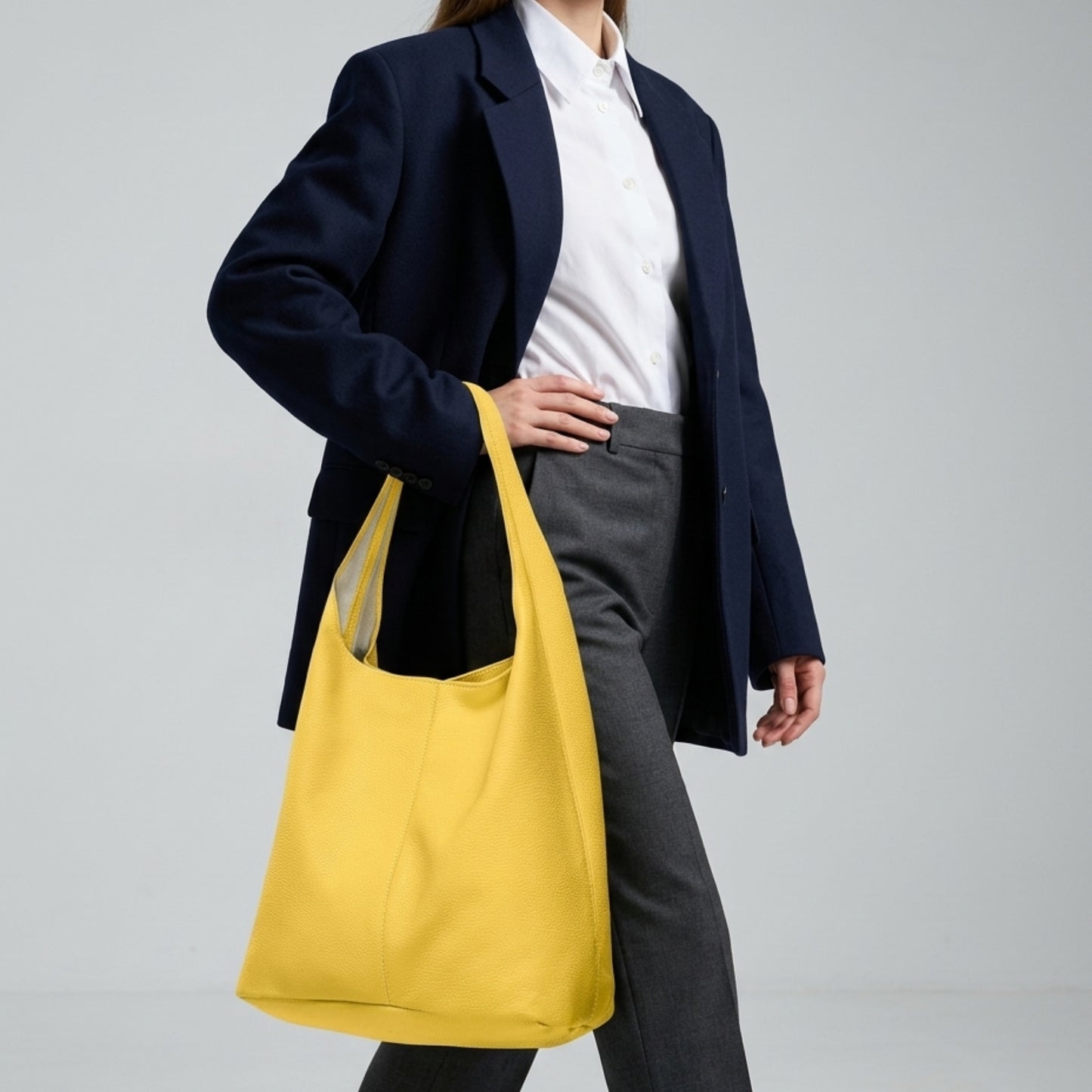 A person in a navy blazer, white shirt, and gray pants walks while carrying an Italian hobo bag on their shoulder against a plain light background.