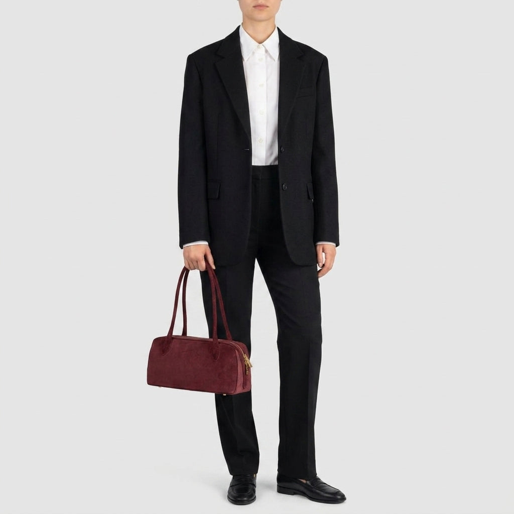 A person in a black suit, white shirt, and black shoes stands holding a structured leather handbag with both handles in their left hand, against a plain light gray background.
