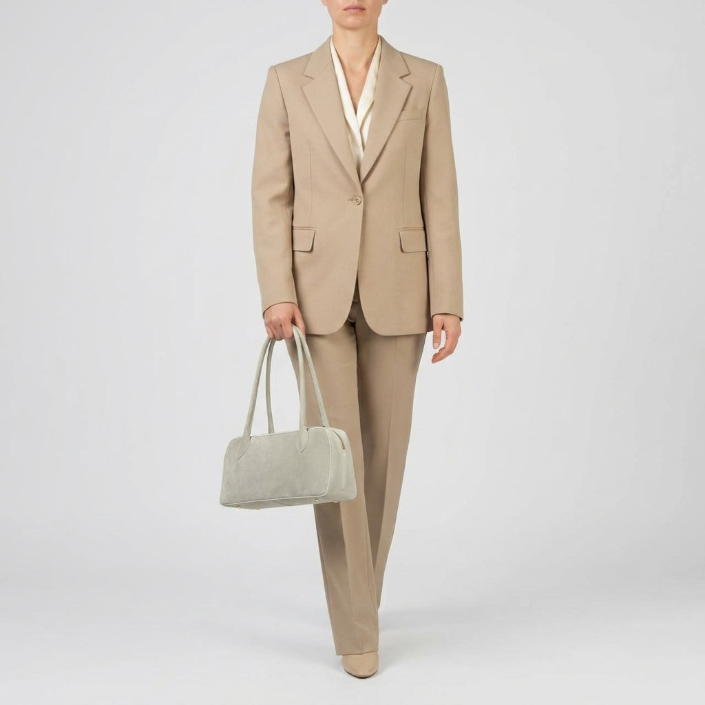 A person wearing a beige pantsuit with a cream blouse stands against a plain background, holding a structured leather handbag in their left hand. Their shoes match the suit, and their face is partially out of frame.