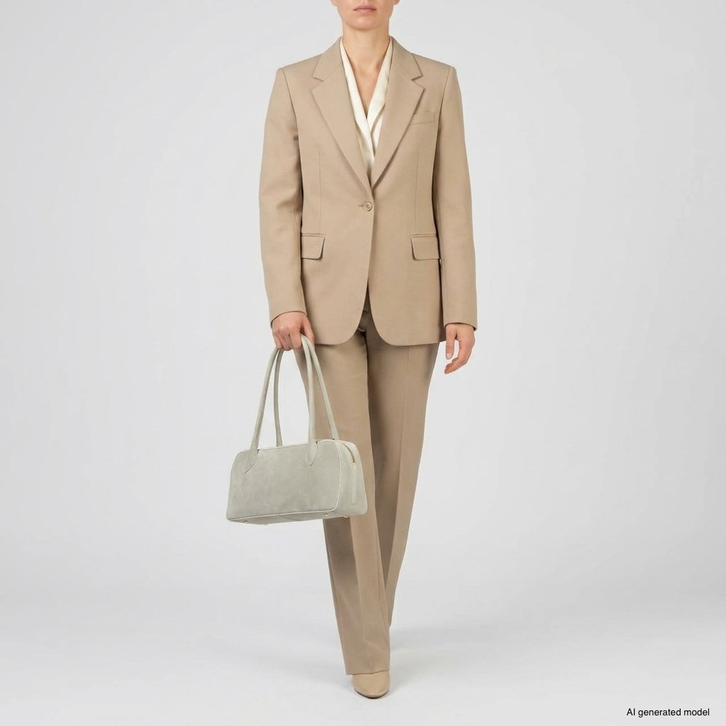 A person wearing a tan suit with a cream blouse holds a structured leather bag in light gray. The outfit includes a tailored blazer and matching pants. The background is plain and light-colored.