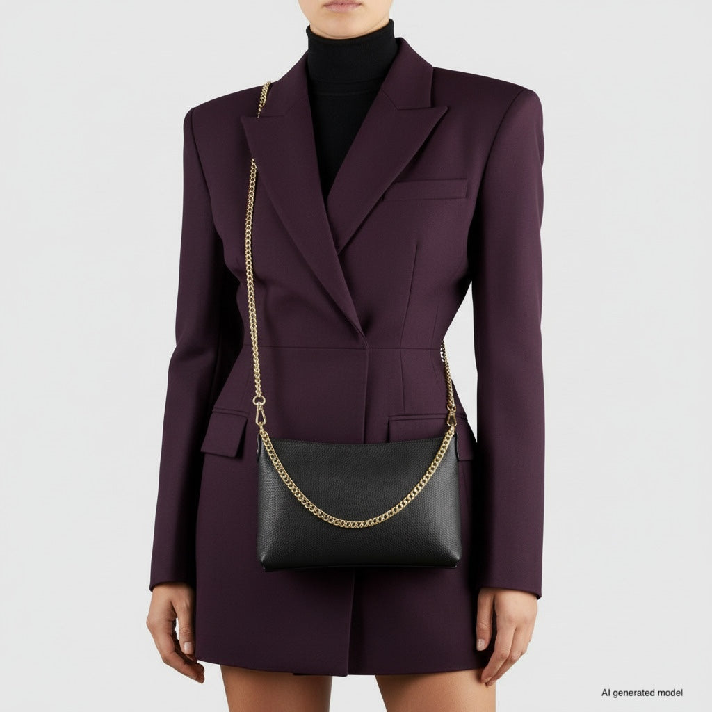 A woman in a dark purple blazer dress holds a small structured handbag with a gold chain shoulder strap. Her head is slightly cropped out of the frame, and the background is plain and light-colored.