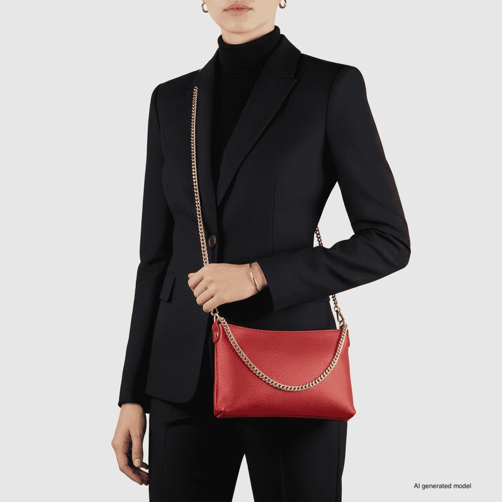 A person in a black suit and turtleneck holds a small structured handbag with a gold chain strap. The Italian leather crossbody purse stands out in red, while the model's face is partially hidden against a plain light grey background.