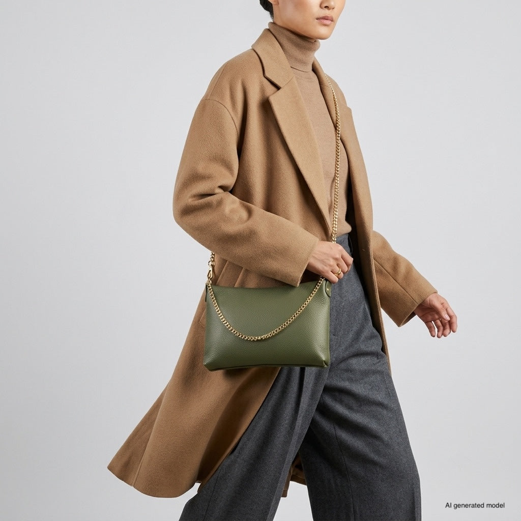 A person wearing a tan coat, brown turtleneck, and gray pants holds a small structured handbag with a gold chain strap, walking against a plain light background.