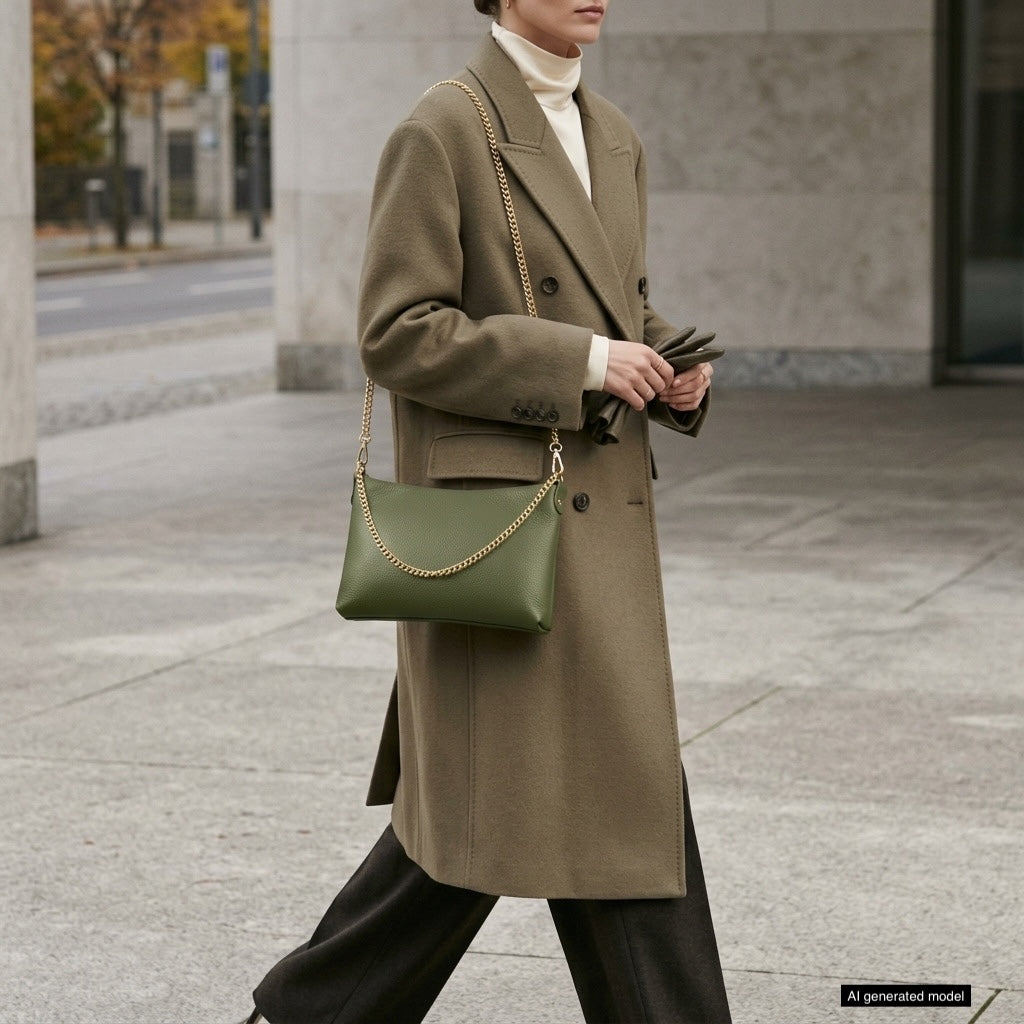 A person wearing a long brown coat, white turtleneck, and dark pants carries a small structured handbag with a gold chain strap while walking outdoors.