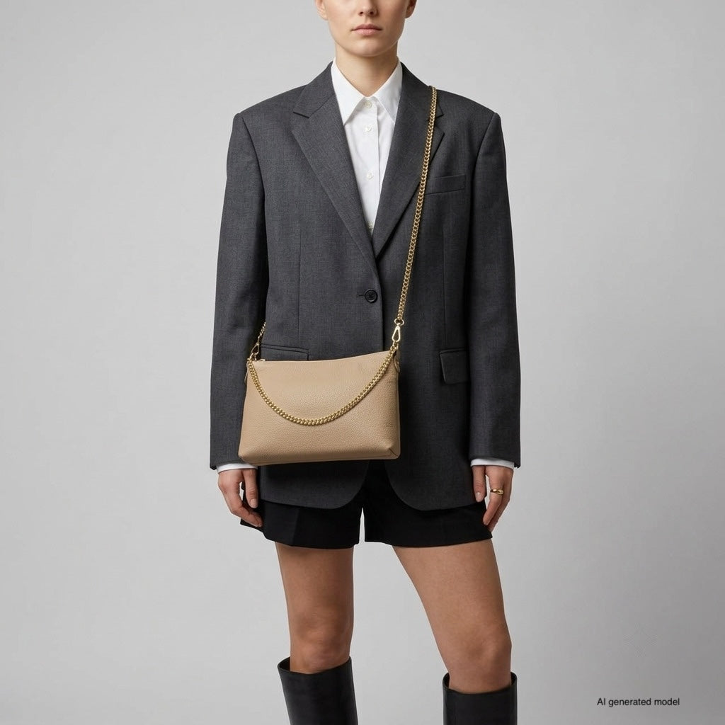 A person wearing a dark gray oversized blazer, white shirt, black shorts, and knee-high black boots stands against a plain background, holding a small structured handbag with a gold chain shoulder bag detail.