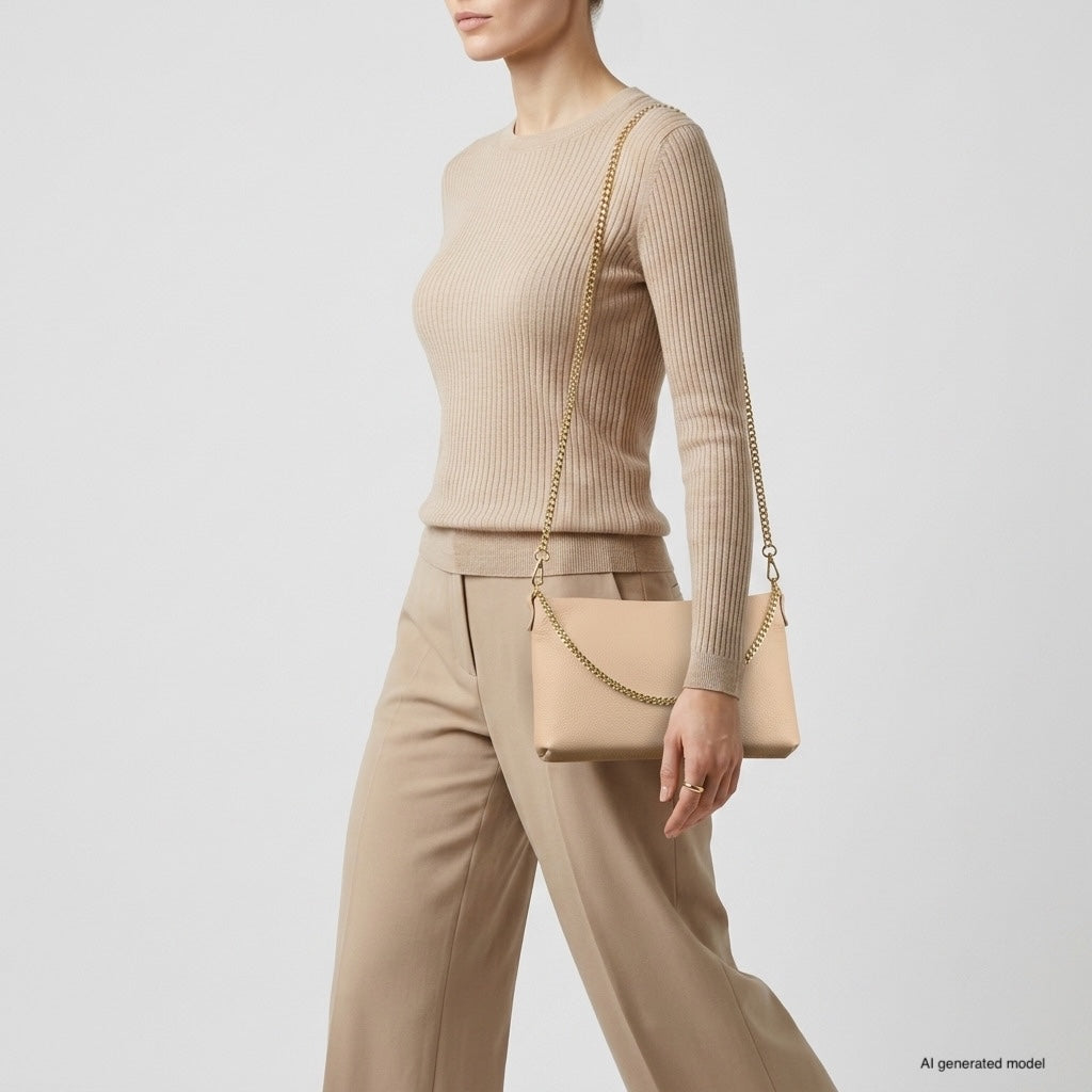 A person wearing a beige ribbed sweater and tan pants carries a small structured handbag with a gold chain shoulder strap over their shoulder against a plain light background.