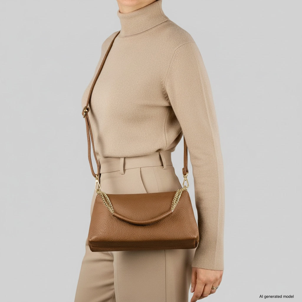 A person wearing a beige turtleneck sweater and matching pants carries a genuine leather crossbody handbag with a gold chain detail, showcasing Italian craftsmanship, against a plain light gray background.