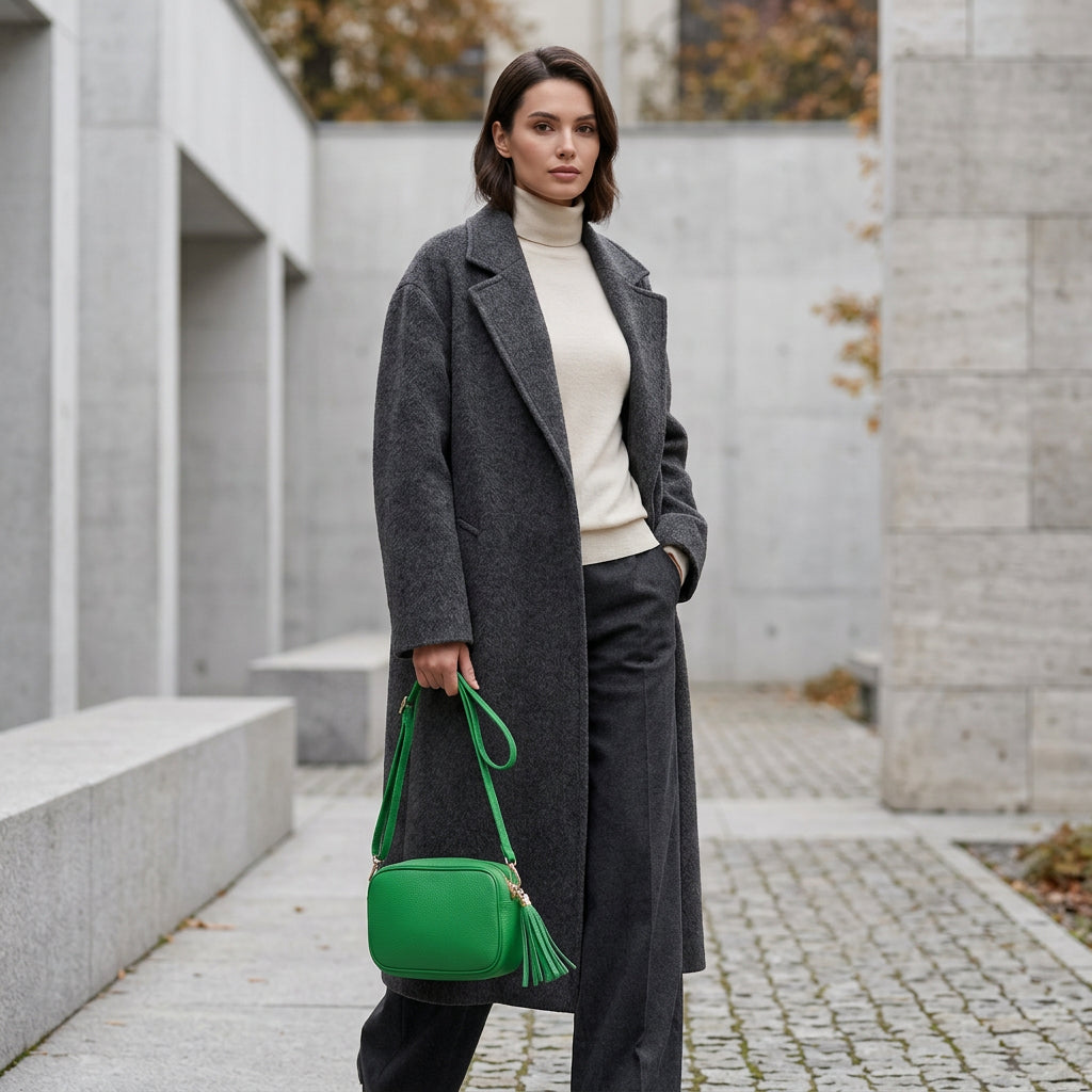 A woman in a long dark gray coat, cream turtleneck, and matching pants stands outdoors on a stone pathway, holding a women’s crossbody bag handcrafted in Italy. The background features modern concrete architecture and autumn foliage.