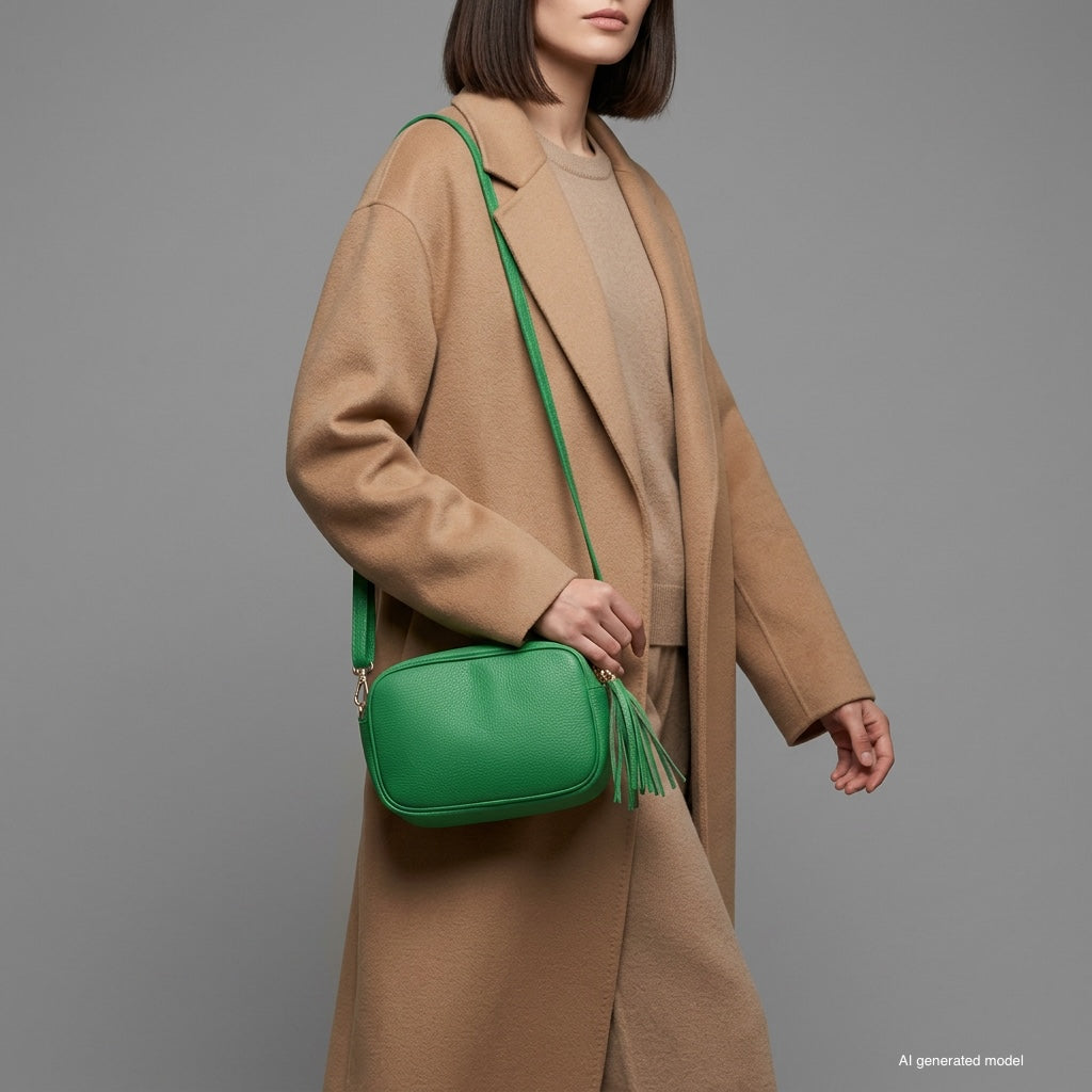 A person wearing a long beige coat and tan outfit carries a bright green handcrafted women’s bag with a tassel. The background is plain gray. The person's face is not visible.