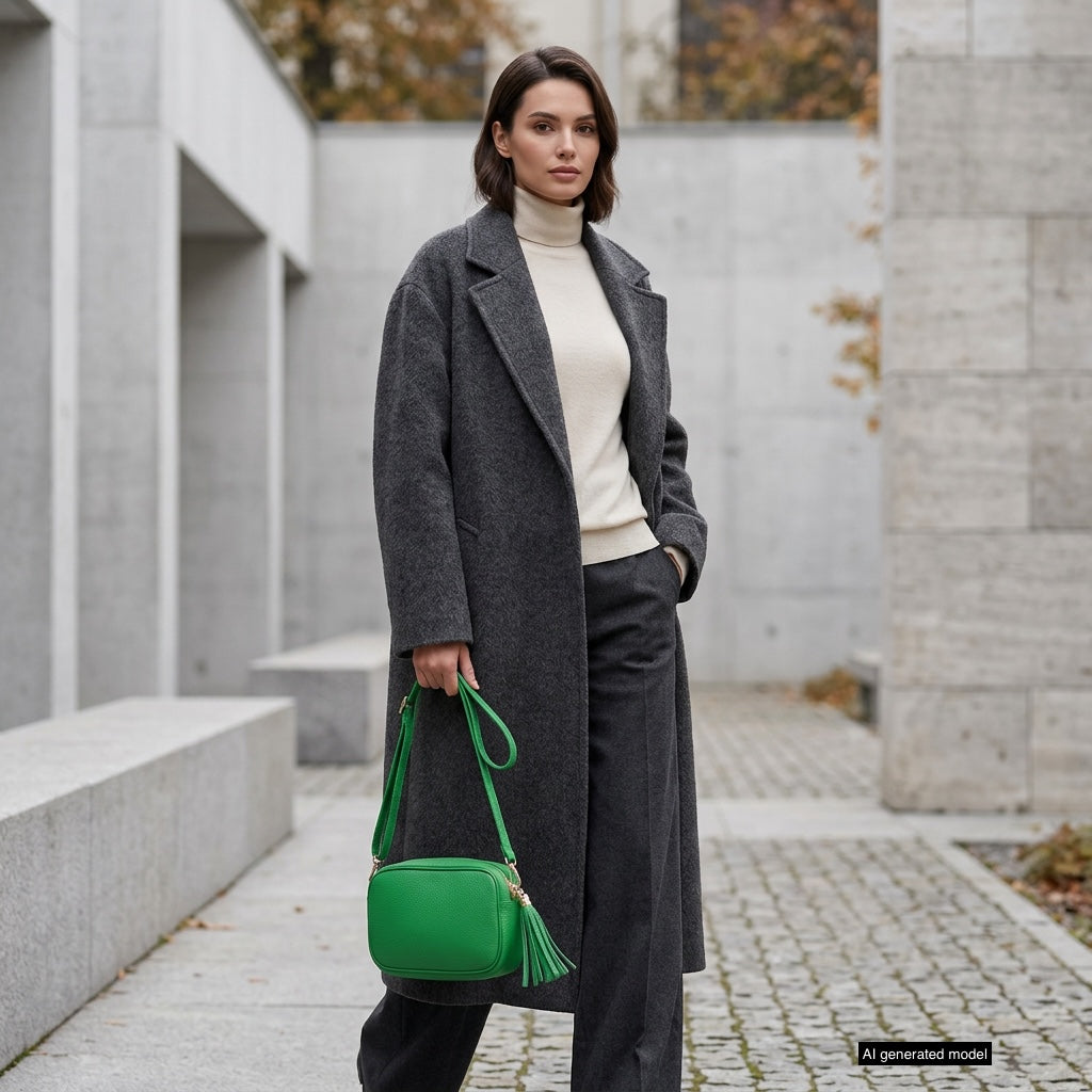 A woman in a long dark gray coat, cream turtleneck, and dark trousers stands outdoors on a stone path, holding a bright green Italian leather crossbody bag. Modern concrete buildings and autumn trees are in the background.