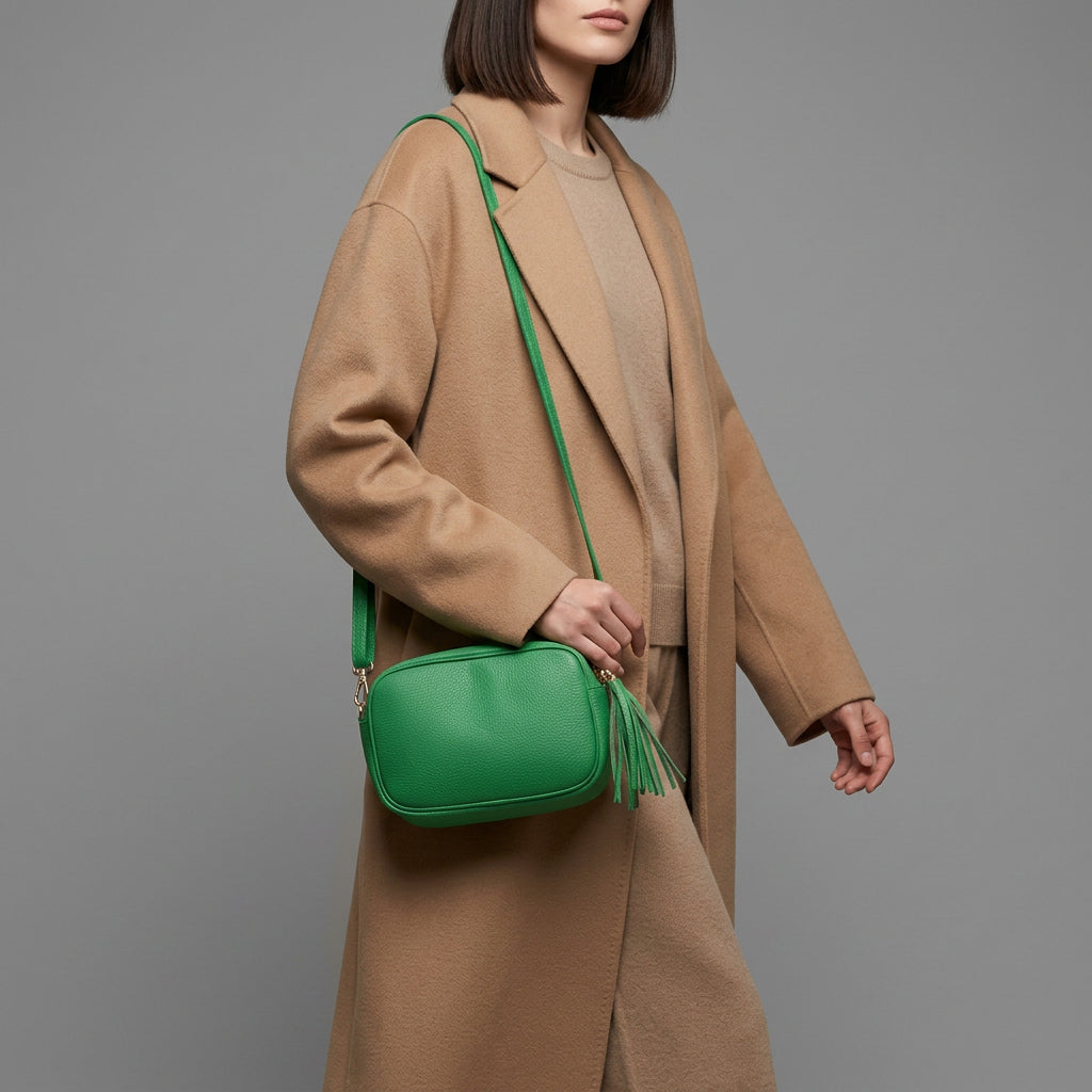 A woman wearing a long, beige coat and tan outfit carries a bright green women’s crossbody bag handcrafted in Italy with a tassel, standing against a plain gray background. Her face is partially out of frame.