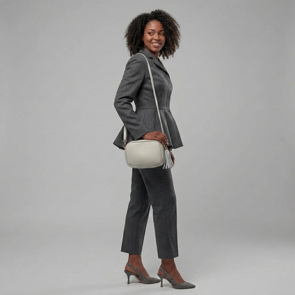 A woman in a grey suit, grey heels, and a white women’s crossbody bag handcrafted in Italy stands smiling against a plain light grey background.