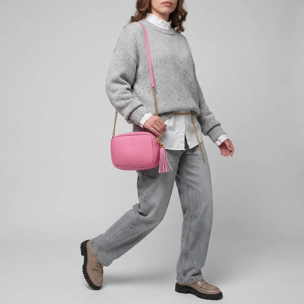 A person wearing a loose grey sweater, white shirt, grey jeans, and beige shoes carries a women’s crossbody bag handcrafted in Italy with a pink tassel and gold chain strap against a plain light background.