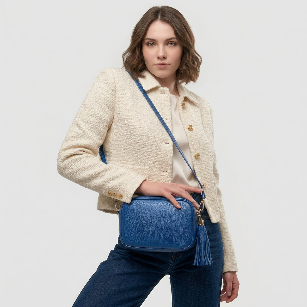 A woman in a cream textured jacket and dark jeans poses with a blue women’s crossbody bag handcrafted in Italy, featuring a large tassel, against a plain light background.