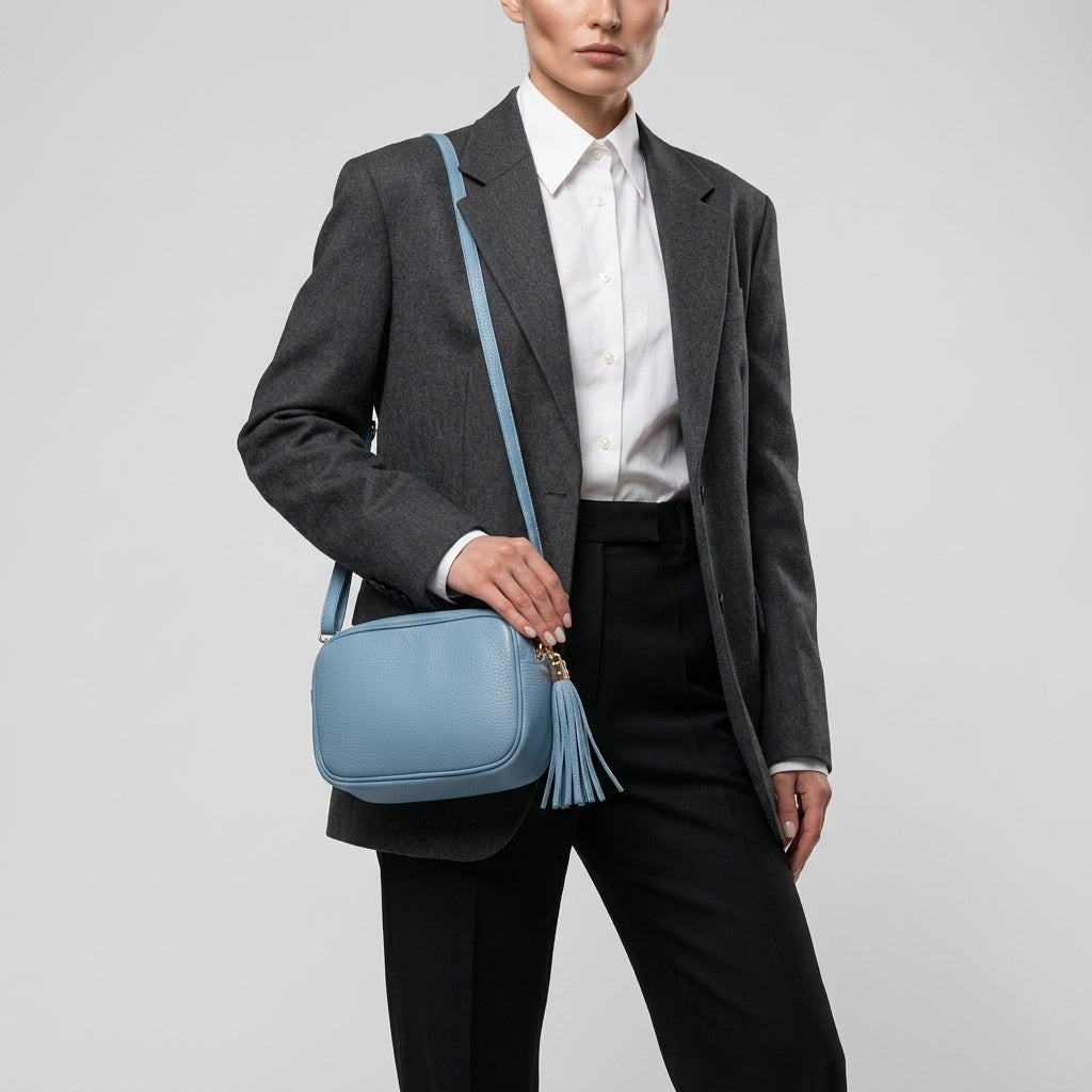 A person wearing a dark gray blazer, white dress shirt, and black pants holds a women’s crossbody bag made from Italian calf leather with a tassel against a plain light gray background.