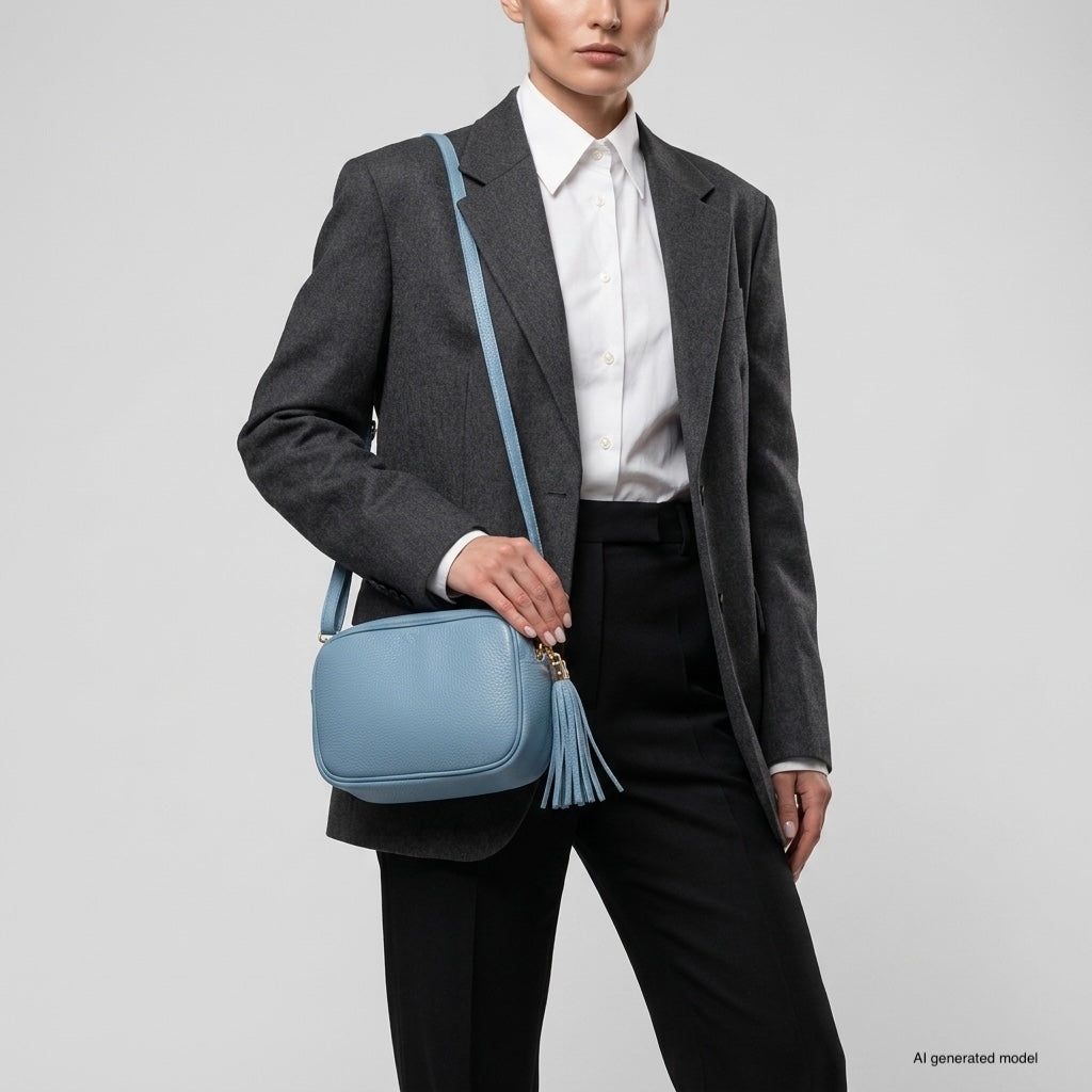 A person wearing a dark gray blazer, white shirt, and black pants holds an Italian leather crossbody bag with a tassel detail. The background is plain and light-colored.