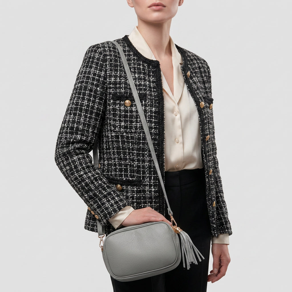 A woman in a black and white tweed jacket, cream blouse, and black pants holds a women’s crossbody bag handcrafted in Italy from fine Italian calf leather, featuring a tassel detail against a plain light background.