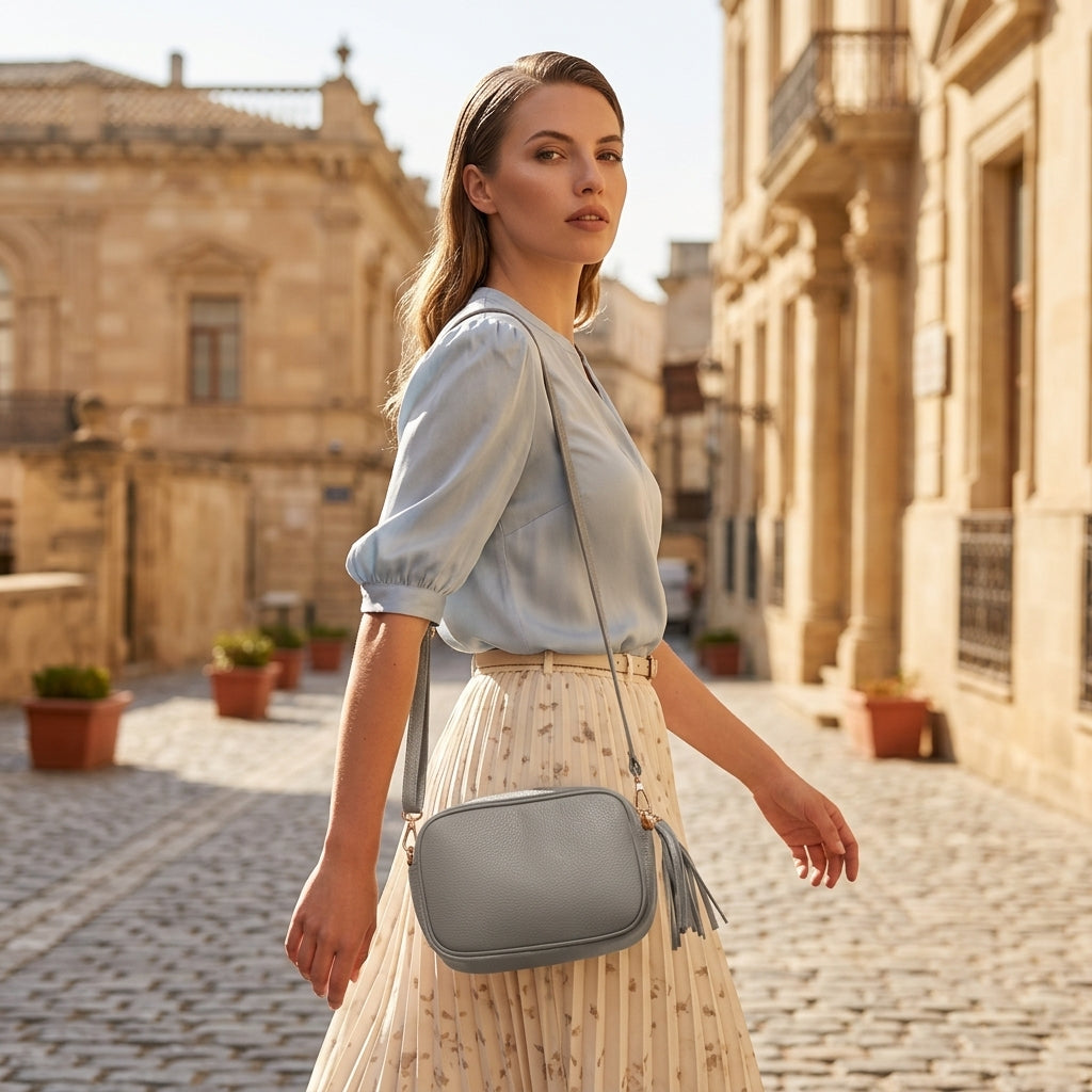 A woman in a light blue blouse and floral skirt walks on a cobblestone street with historic buildings, carrying a women’s crossbody bag handcrafted in Italy, and looking over her shoulder.