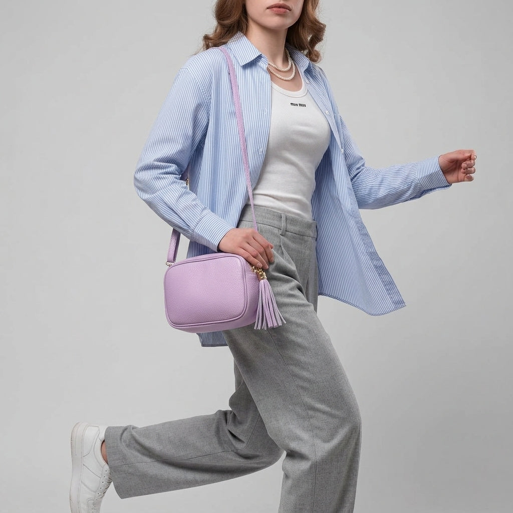A woman in gray pants, a white top, and a blue striped shirt walks with a women’s crossbody bag, handcrafted in Italy from Italian calf leather. She wears white sneakers and layered necklaces against a plain gray background.