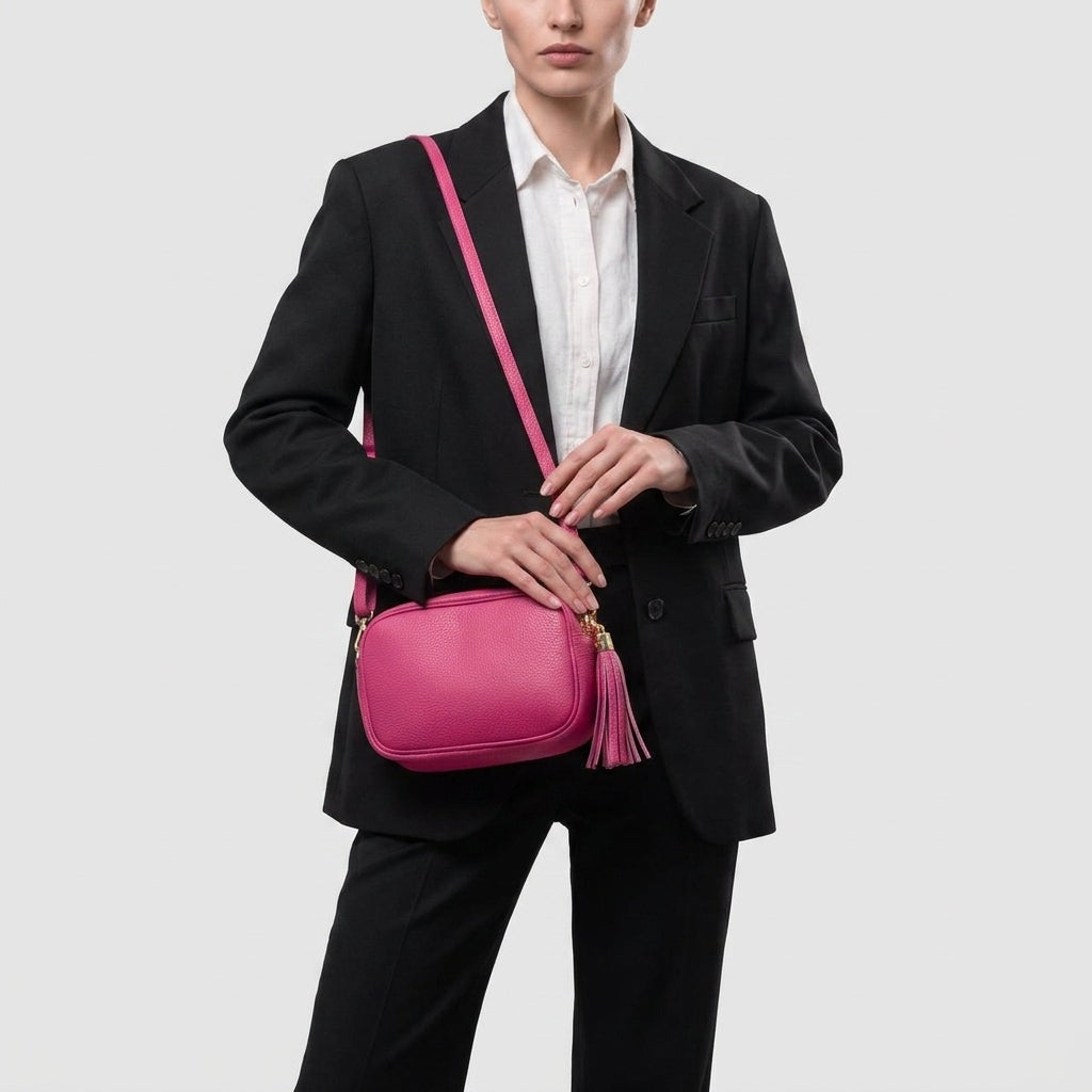 A person in a black suit and white shirt holds a bright pink women’s crossbody bag with a tassel, handcrafted in Italy, standing against a plain light gray background.