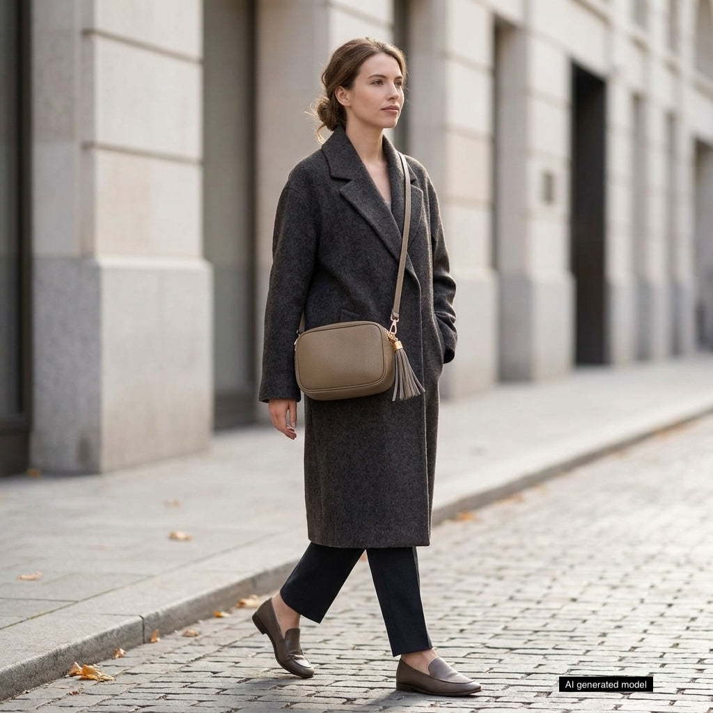 A woman in a long dark coat, black pants, and brown loafers walks on a city sidewalk. She carries an Italian leather crossbody bag and appears calm, with buildings lining the street in the background.