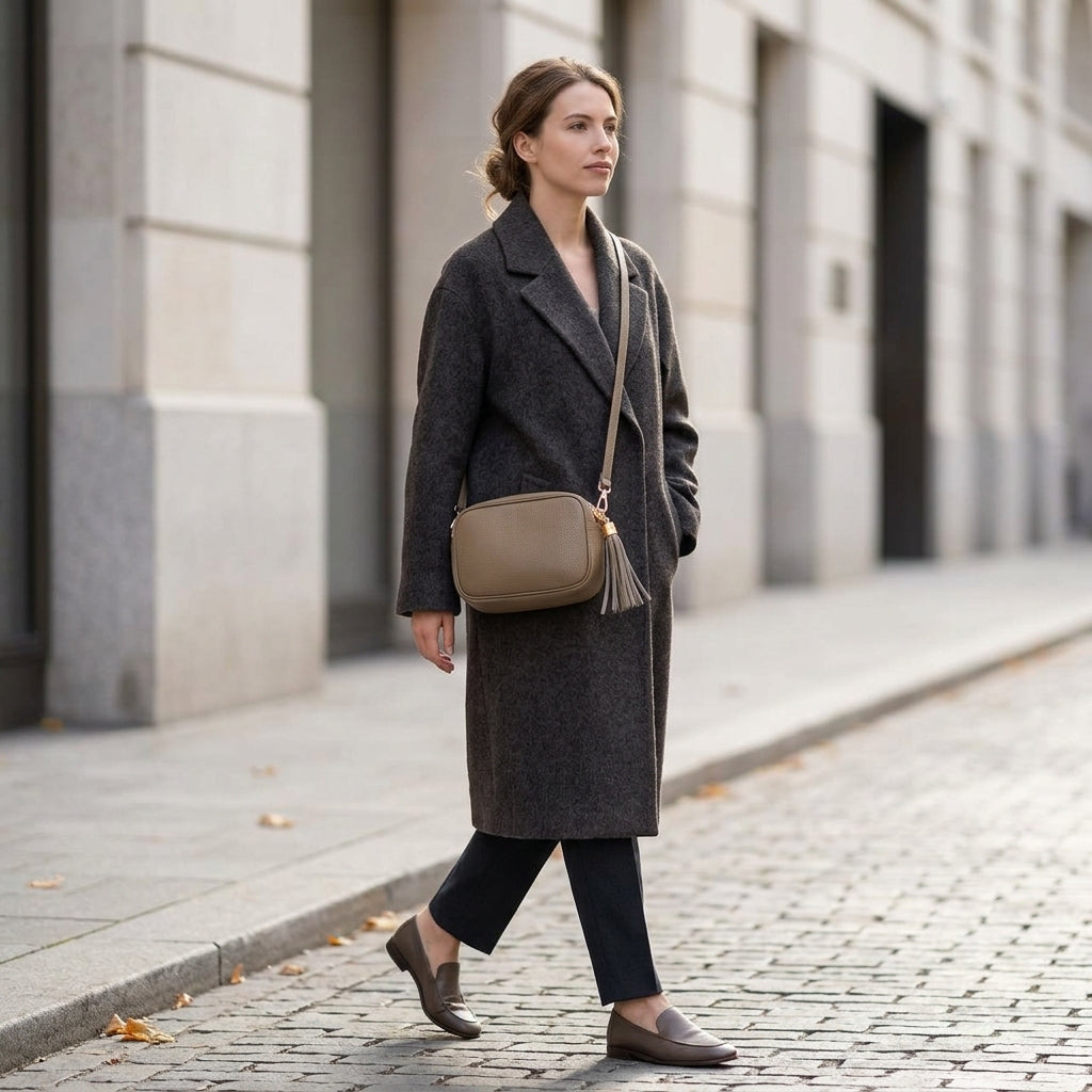 A woman in a long dark coat, black pants, and brown loafers walks on a cobblestone sidewalk with a beige women’s crossbody bag handcrafted in Italy. She passes light stone buildings as fallen leaves scatter across the ground.