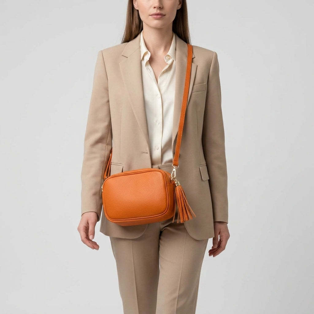 A woman wearing a beige suit and white blouse carries an orange women’s crossbody bag, handcrafted in Italy from fine Italian calf leather, with a tassel detail, standing against a light gray background.