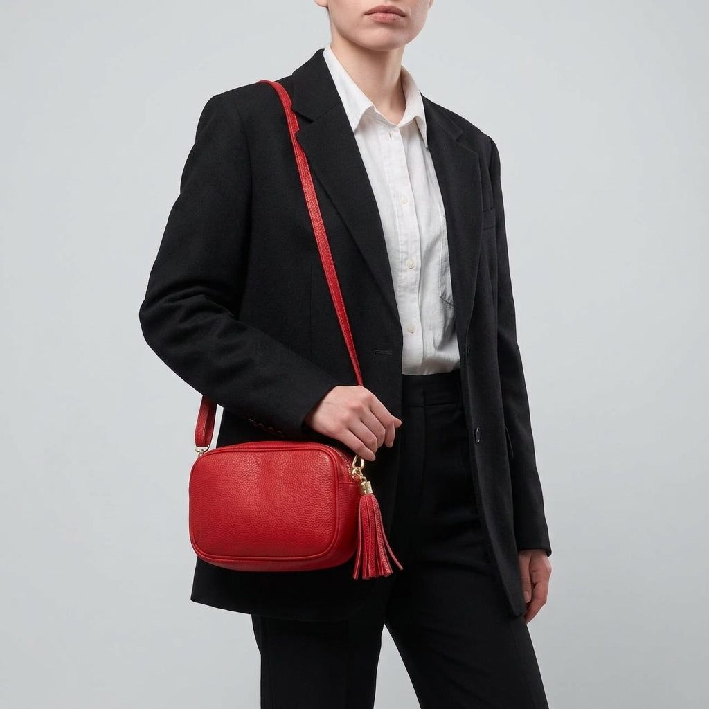 A person wearing a black blazer and white shirt holds a small red women’s crossbody bag handcrafted in Italy, with a tassel, standing against a plain light gray background.