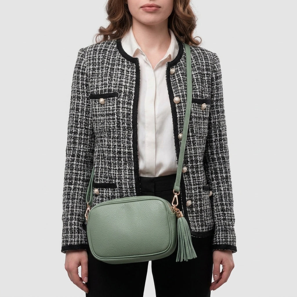 A woman wearing a black and white tweed jacket, white blouse, and black pants stands with a light green women’s crossbody bag featuring a tassel detail, handcrafted in Italy from luxurious Italian calf leather.