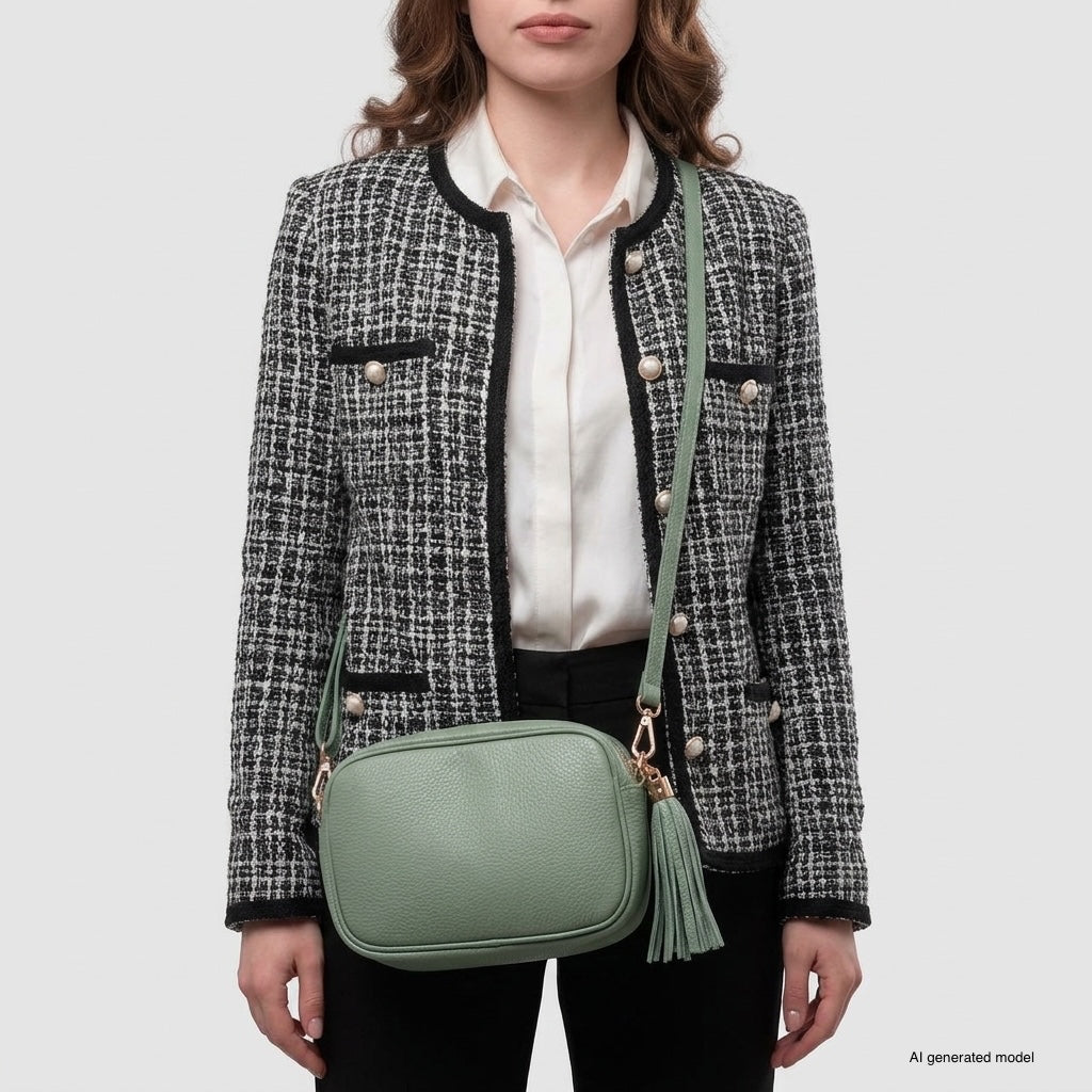 A woman in a black and white tweed jacket and black pants stands with an Italian leather crossbody bag featuring a tassel, posed against a plain gray background.
