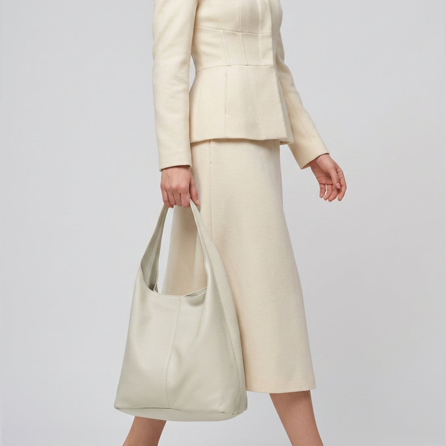 A person in a cream-colored suit and skirt walks while holding a large, handmade shoulder purse by their side against a plain background. Their head is not visible in the image.