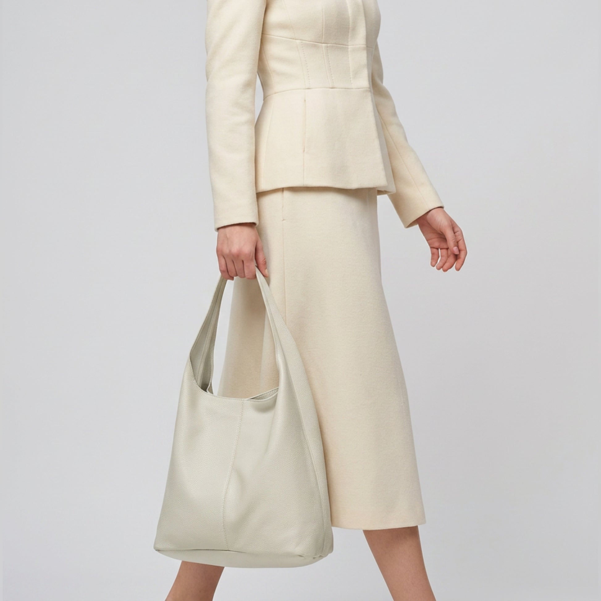 A person in a cream-colored suit and skirt walks while holding a large, handmade shoulder purse by their side against a plain background. Their head is not visible in the image.