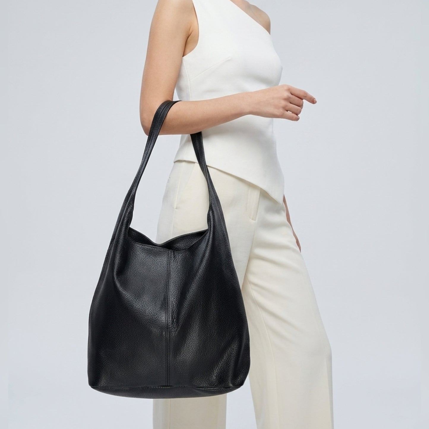 A woman in a sleeveless white top and white pants carries a genuine calf leather Italian hobo bag over her shoulder against a plain light background.