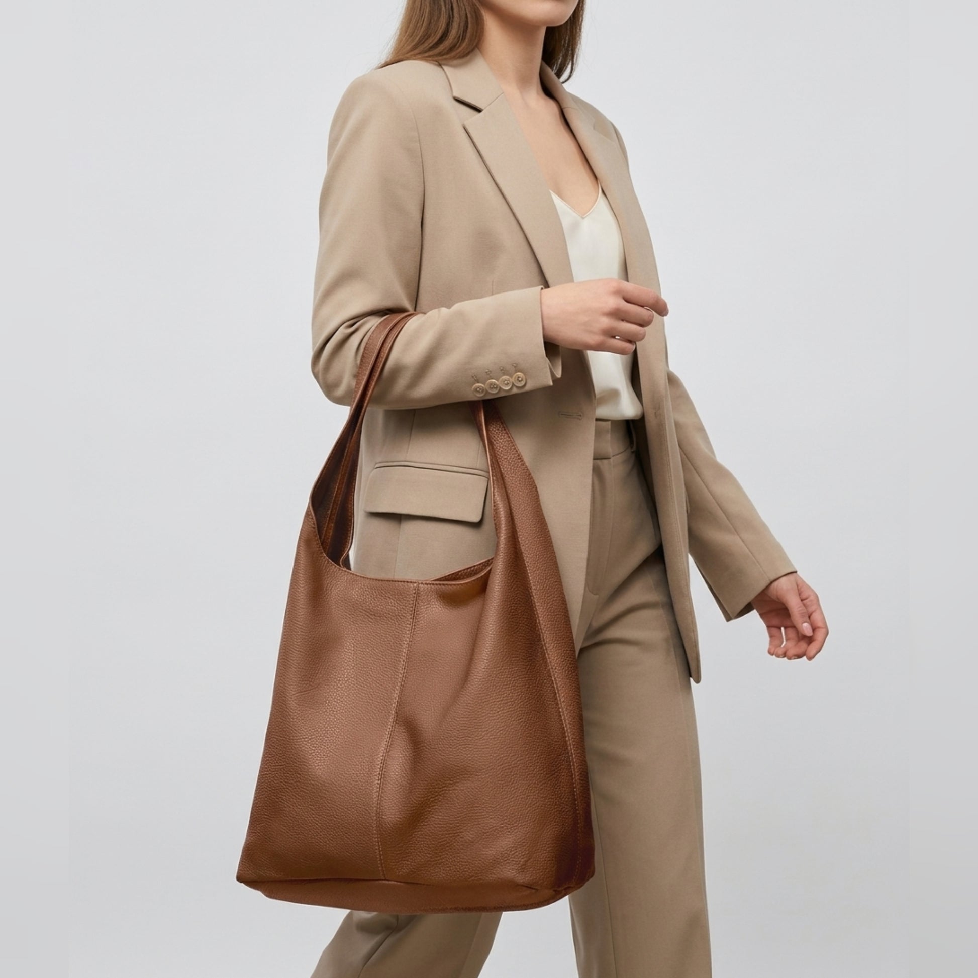 A woman in a beige suit and white top carries a handmade shoulder purse, its rich brown leather slung over her shoulder as she walks against a plain light background.