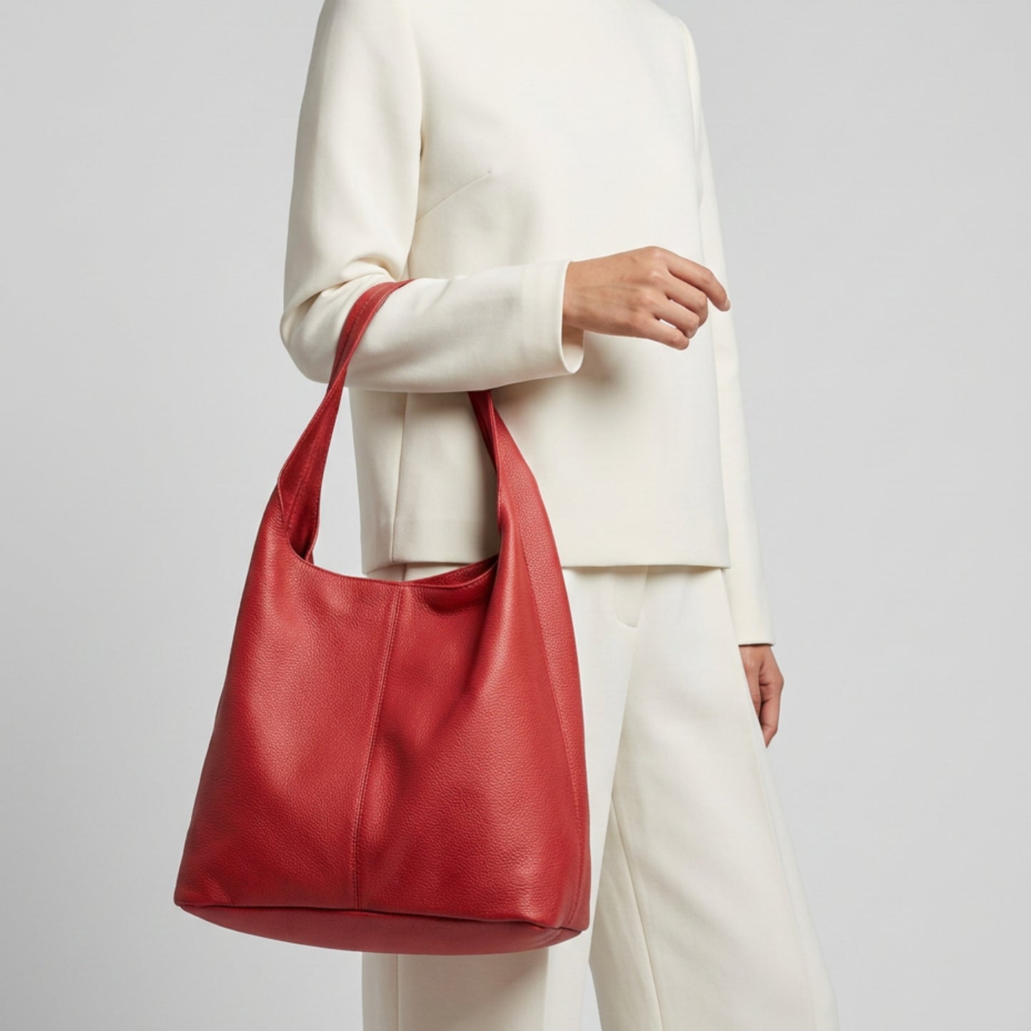 A person wearing an off-white long-sleeve top and pants holds a large, textured Italian leather hobo bag by its handle against a plain light background.