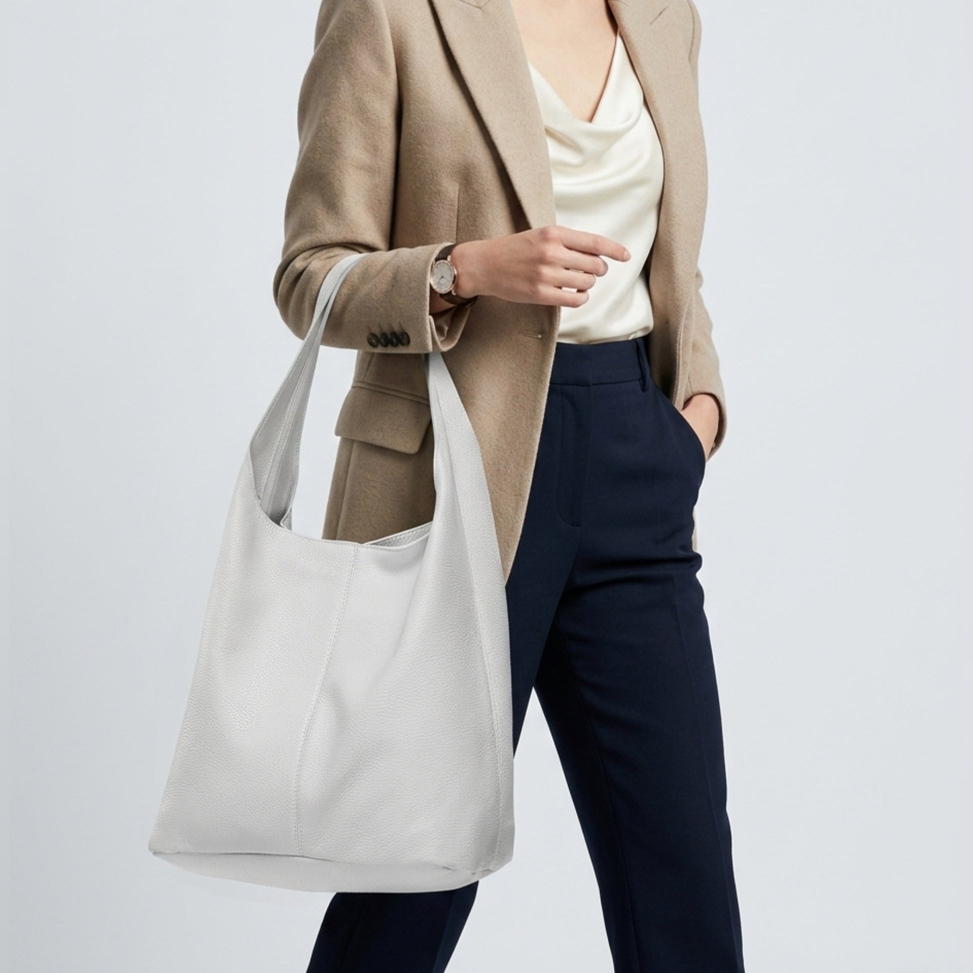 A woman wearing a beige blazer, cream blouse, and navy pants carries a handmade shoulder purse on her shoulder. Only her upper body is visible against a plain light background.