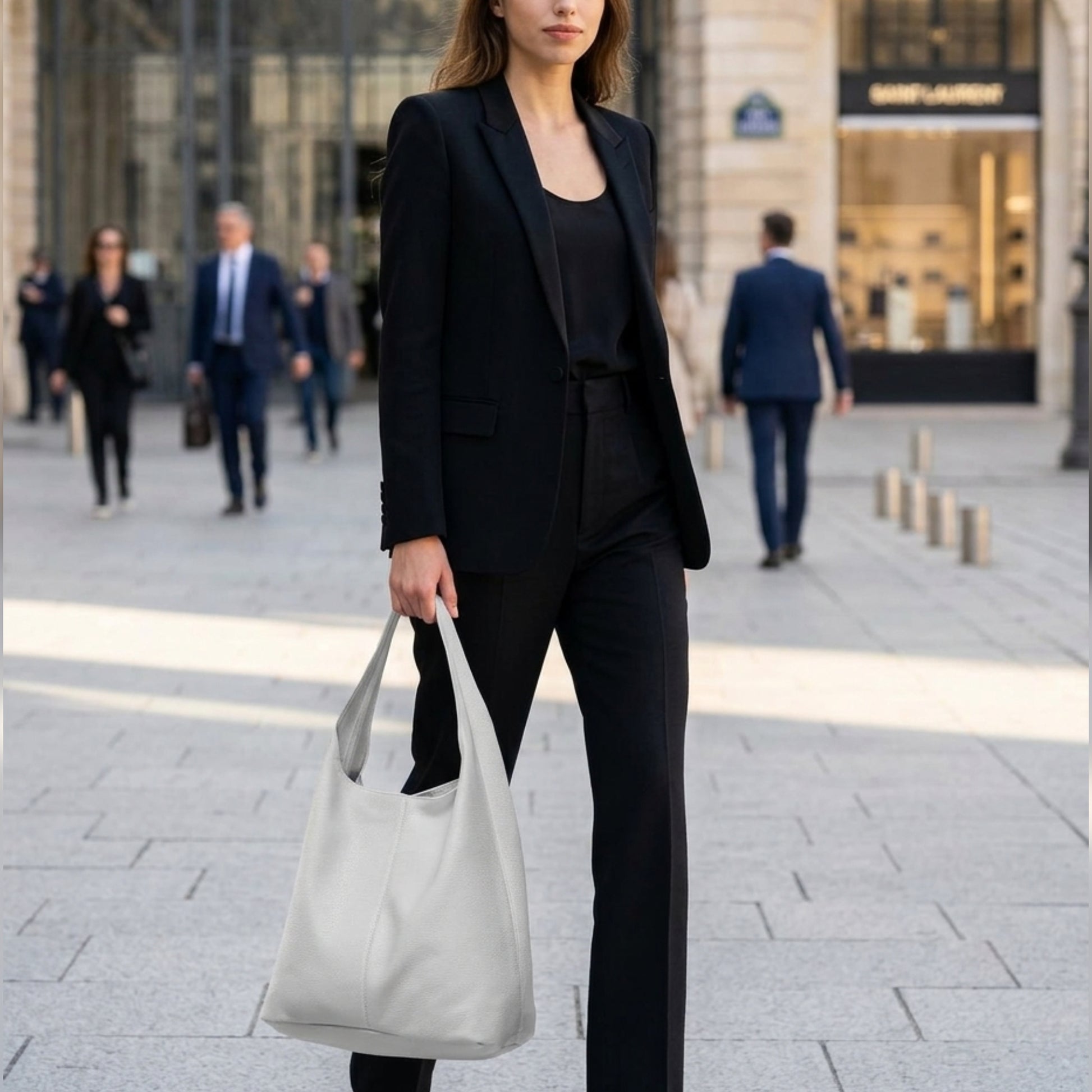 A woman wearing a black suit and carrying an Italian hobo bag walks confidently down a city street, with blurred pedestrians and an upscale store in the background.