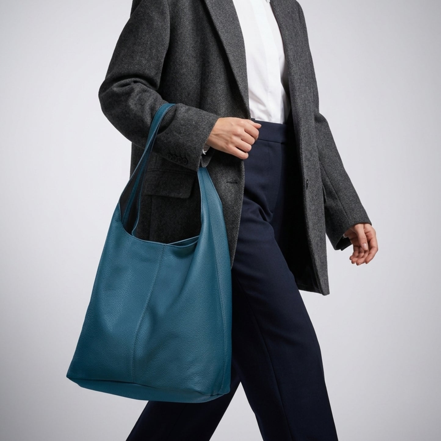 A person wearing a grey blazer, white shirt, and navy trousers walks while carrying a genuine calf leather Italian hobo bag on their shoulder against a plain light background.