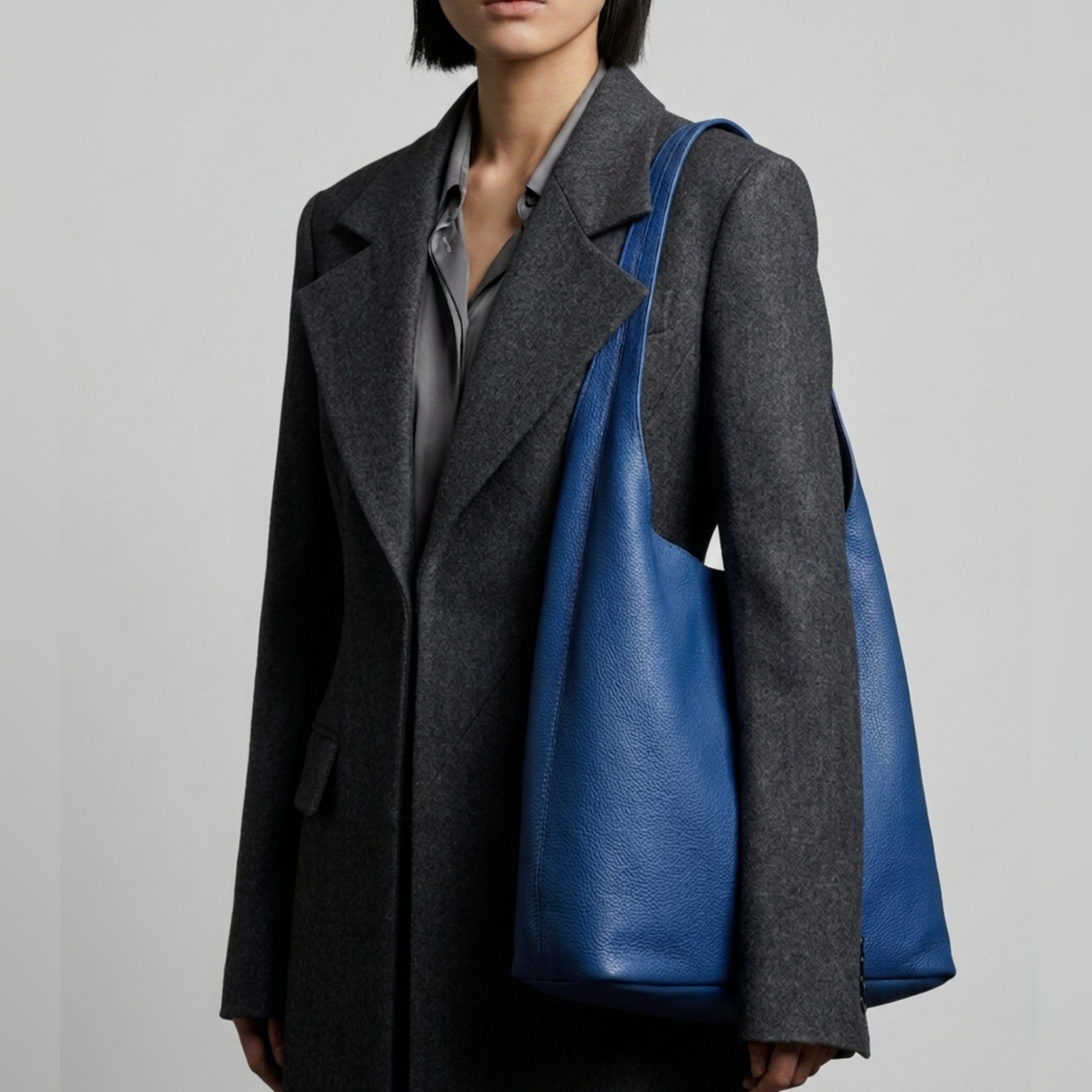 A person wearing a gray coat and shirt carries a large blue Italian hobo bag made from genuine calf leather. The background is plain light gray, and the person's face is partially out of frame.