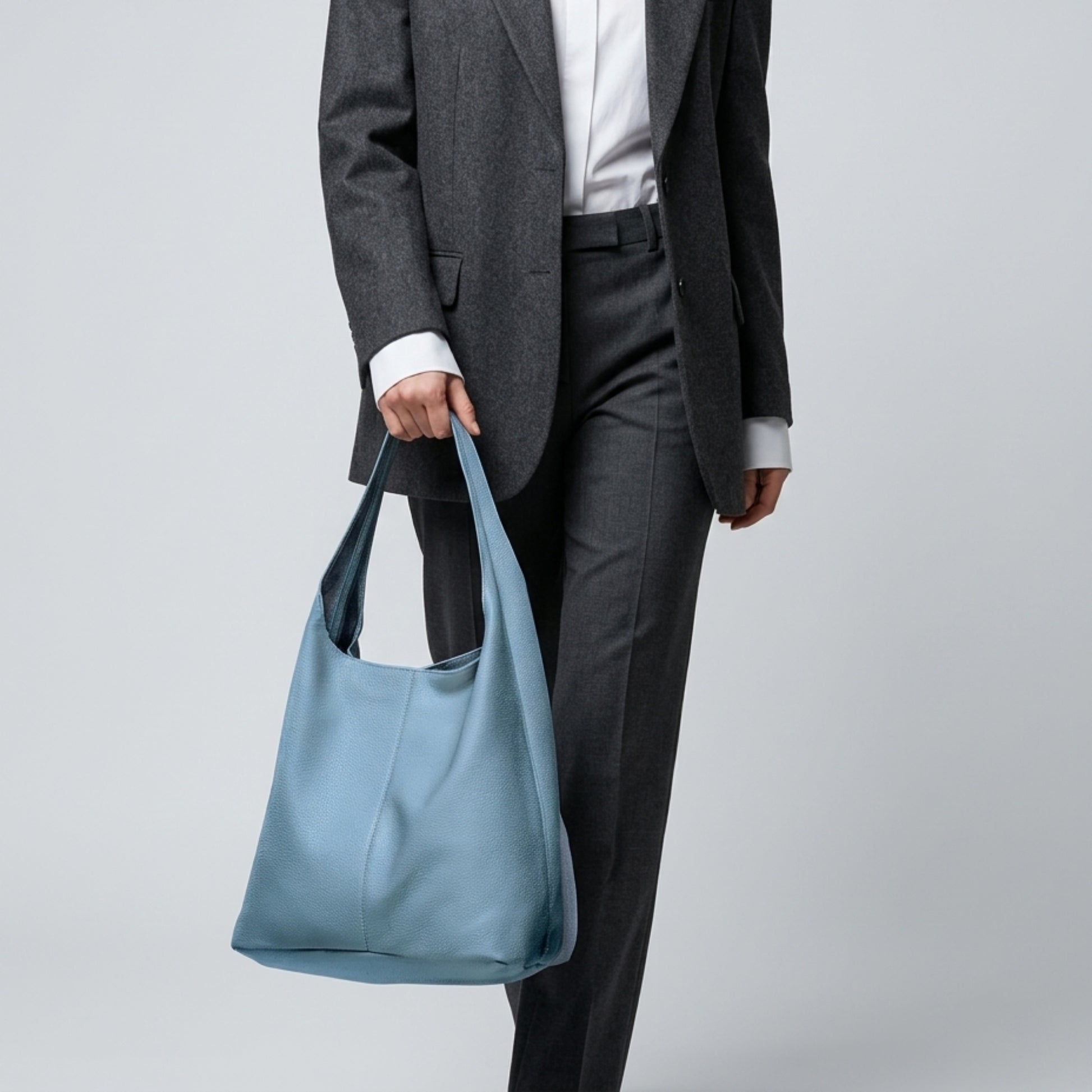 A person in a dark gray suit and white shirt holds a large, genuine calf leather handbag by its strap. The light blue Italian hobo bag stands out against the plain light gray background.