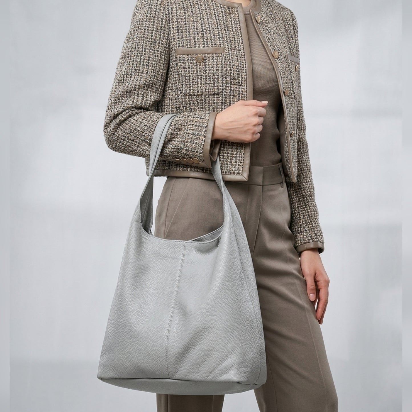 A woman wearing a textured beige jacket and taupe pants holds a large, handmade shoulder purse over her shoulder. The background is plain and neutral.