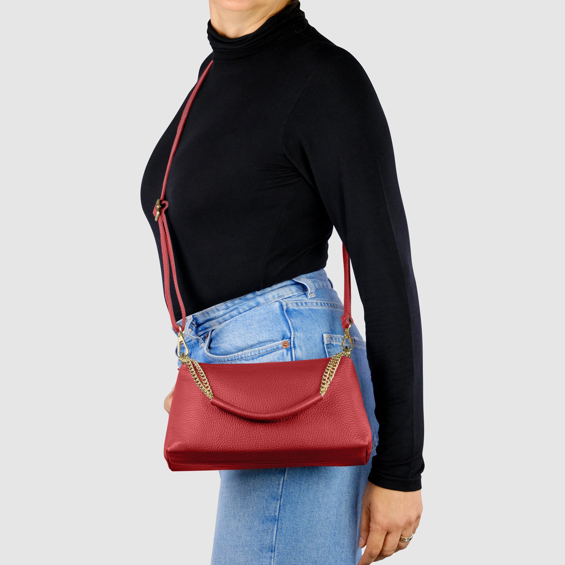 A woman wearing a black turtleneck and blue jeans carries an elegant Italian leather bag with a gold chain strap over her shoulder against a plain light gray background.