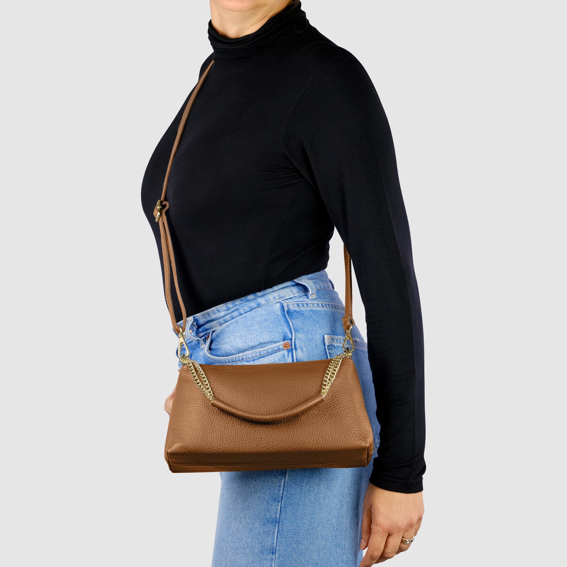 A person wearing a black turtleneck and blue jeans stands sideways, modeling an elegant Italian leather bag with a gold chain detail against a light grey background.