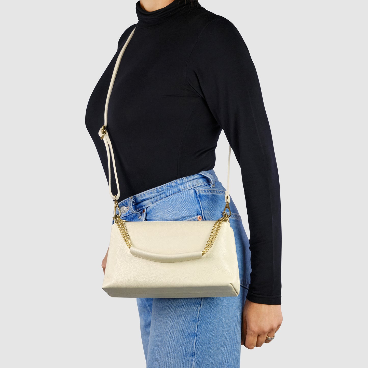 A person wearing a black turtleneck and blue jeans stands sideways, modeling a cream-colored Italian leather bag with a gold chain strap over their shoulder against a plain light background.