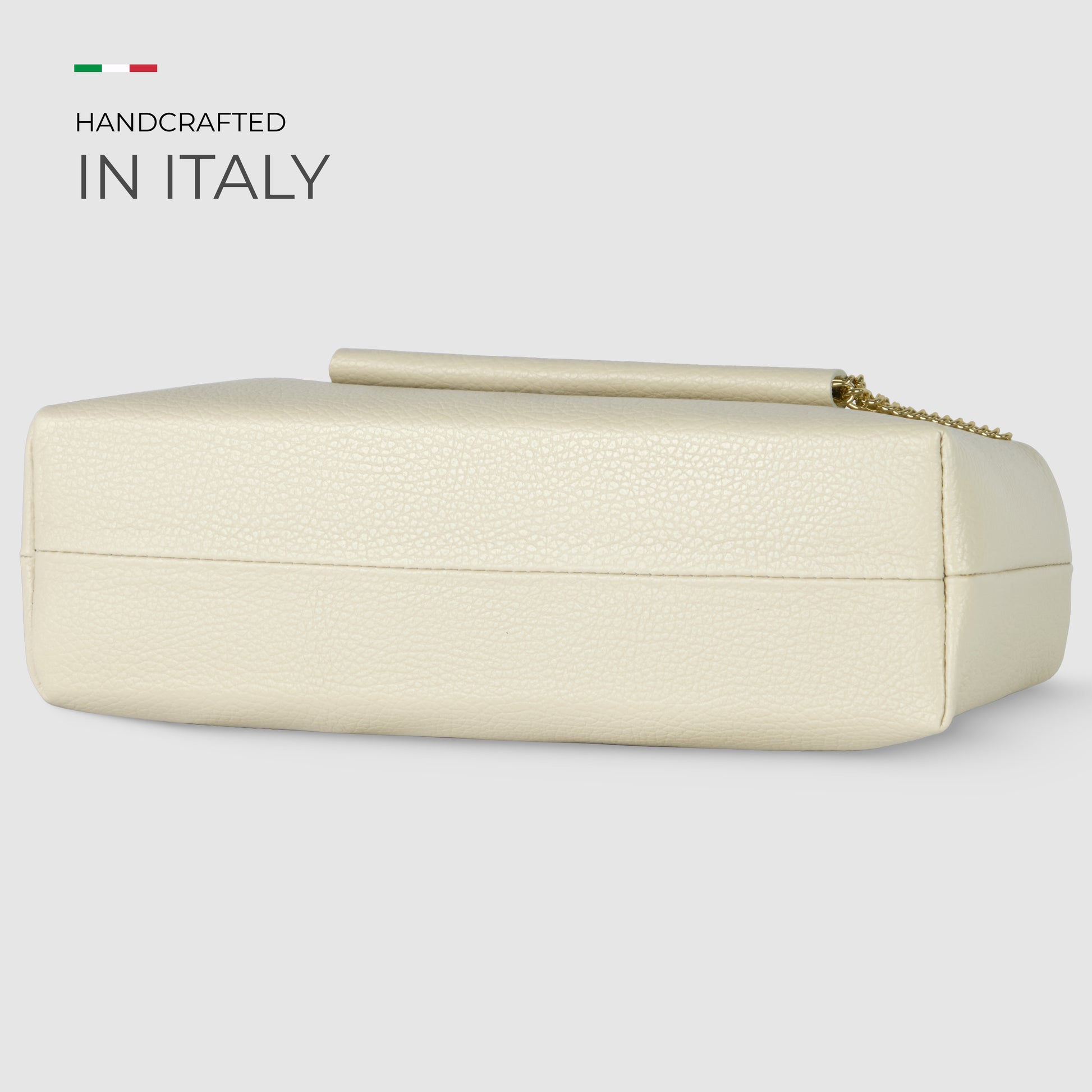 A rectangular, ivory-colored Italian leather bag with a gold chain, displayed on its side. Text reads Handcrafted in Italy next to a small Italian flag.