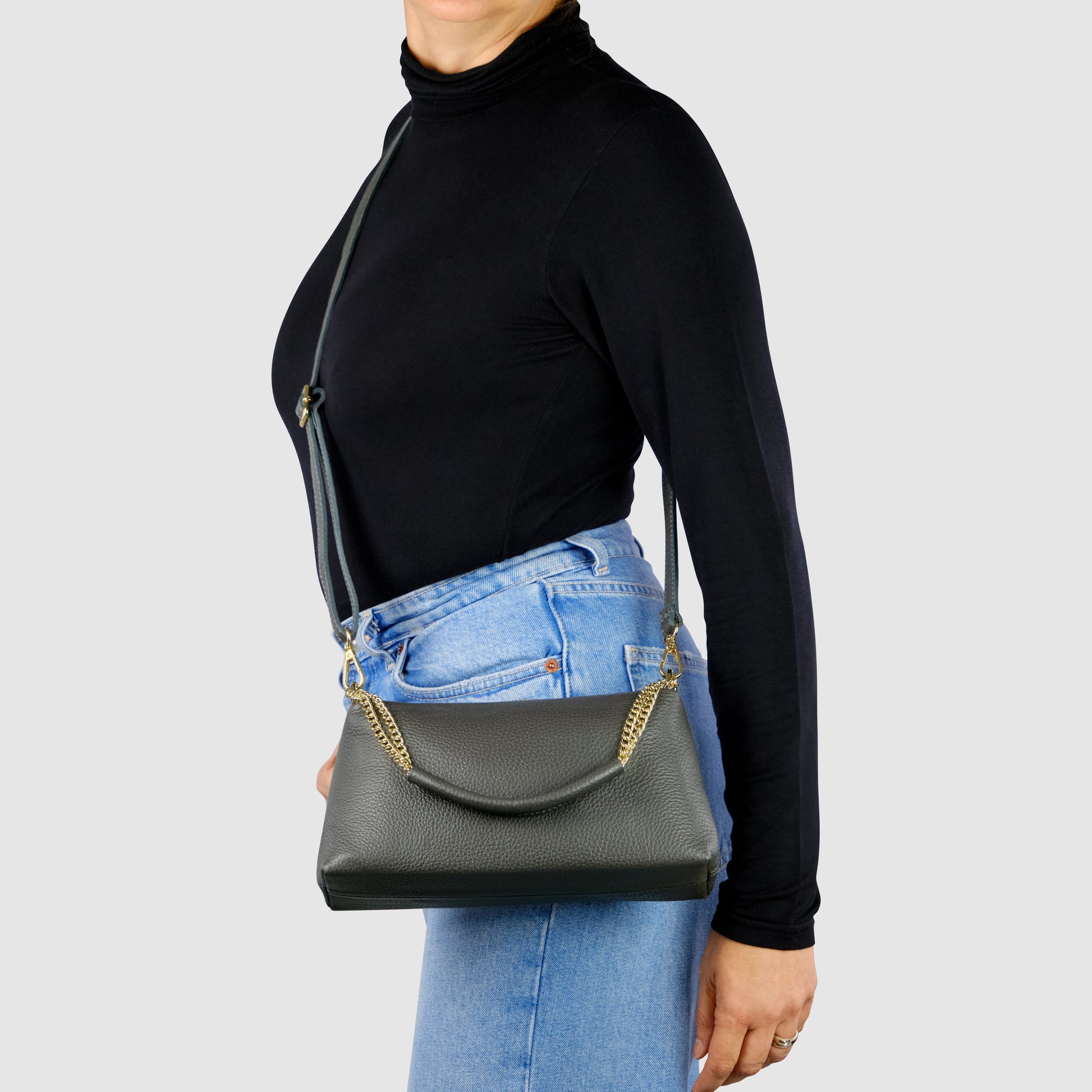 A person wearing a black turtleneck and blue jeans carries a luxury shoulder bag with gold chain details over their shoulder against a plain light gray background.