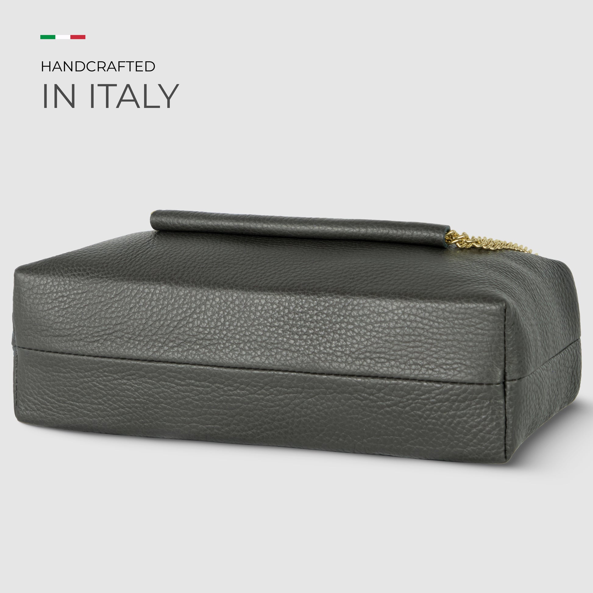 A dark gray, textured Italian leather bag with a gold chain partially visible, displayed on a light gray background. Text above reads “Handcrafted in Italy” with a small Italian flag.