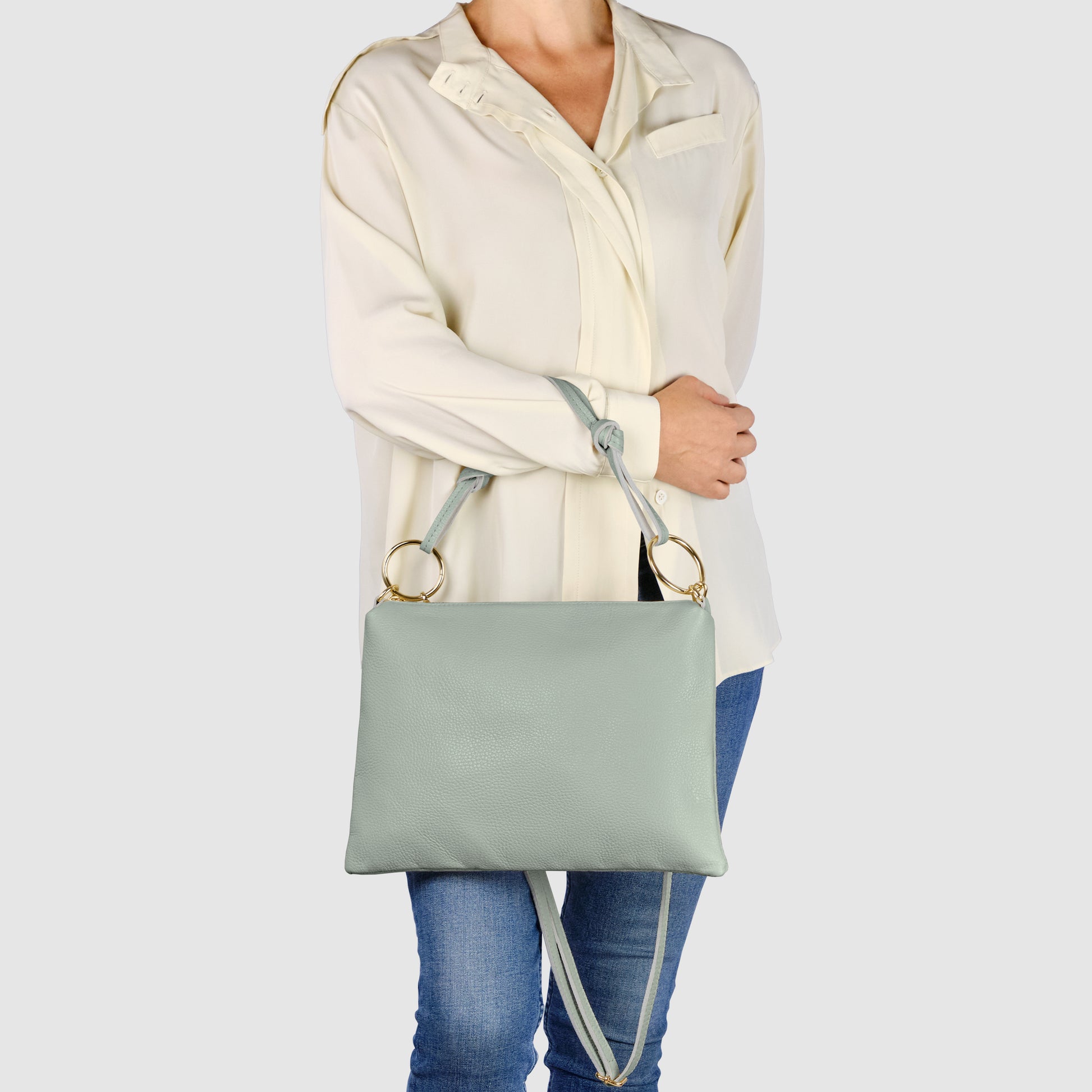 A person wearing a cream-colored blouse and blue jeans holds a structured zippered purse made from Italian leather with gold rings and a crossbody strap against a plain background.